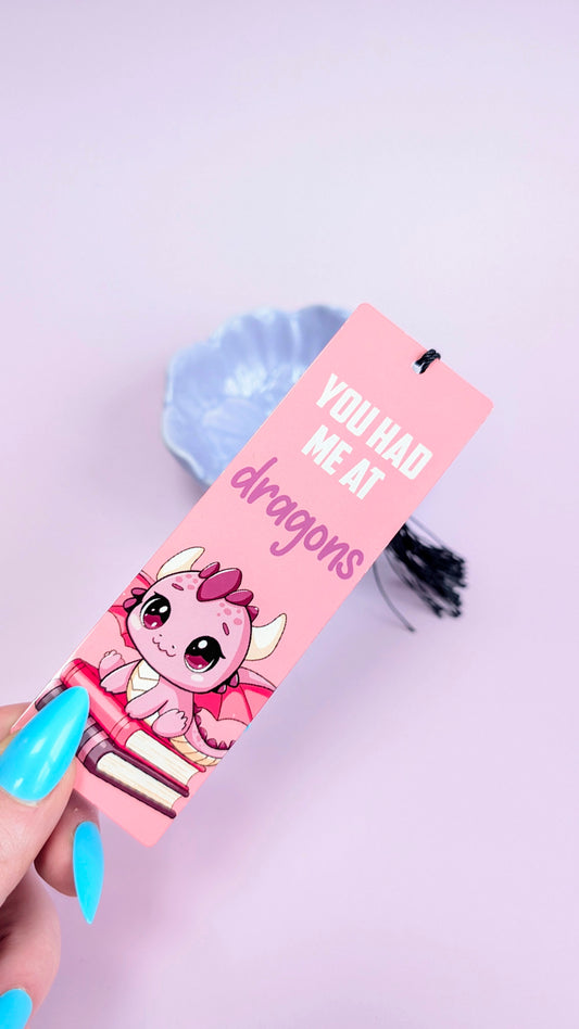 Pink bookmark with a cartoon character and text 'You had me at dragons' held by a hand with blue nail polish on a light purple background.