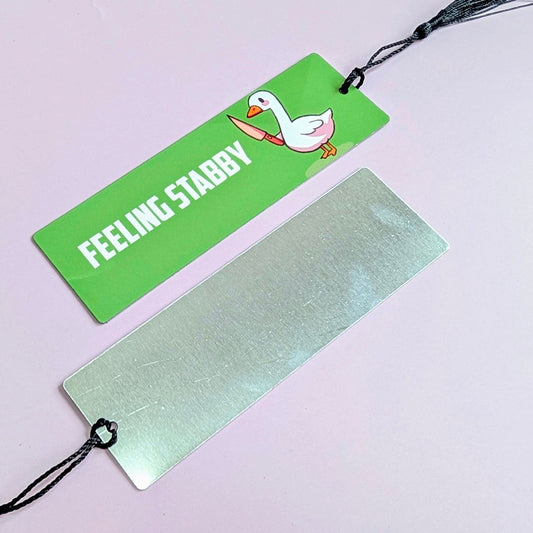 Two bookmarks, one green with a duck illustration and text, the other plain metallic, on a light purple background.