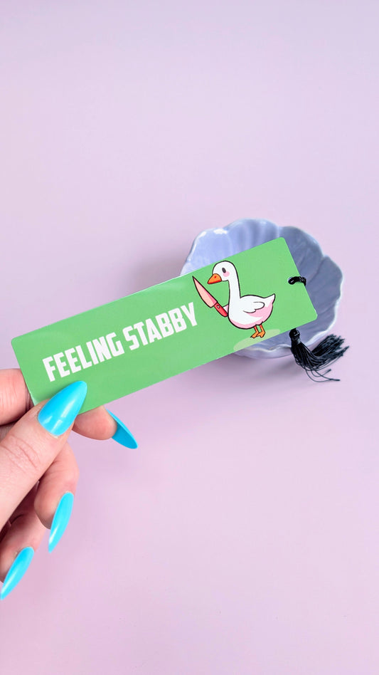 Hand holding a green bookmark with a goose illustration and 'Feeling Stabby' text on a light purple background.