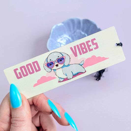 Hand holding a bookmark with a cartoon dog and 'Good Vibes' text against a light purple background